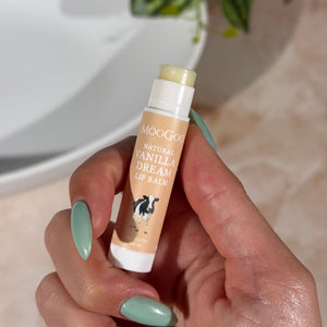 Hand holding a beige tube of MooGoo Natural Vanilla Dream Lip Balm with a plant and bathroom sink in the background.