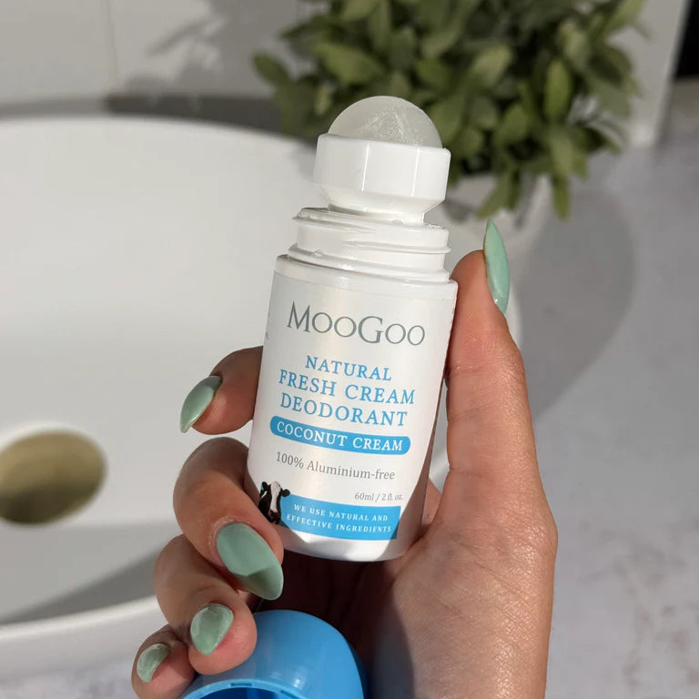 MooGoo Natural Coconut Cream Fresh Cream Deodorant 60ml held by a hand with a tiled background, a plant and bathroom sink.