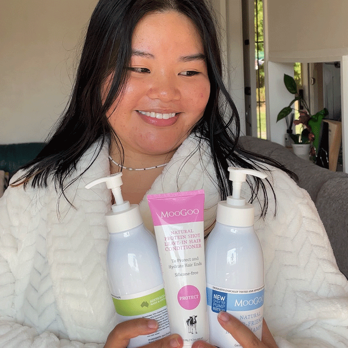 Smiling woman holding MooGoo Protein Shot Leave-In Conditioner and hair care bottles, natural silicone-free conditioning products