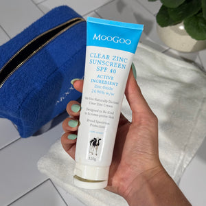 Hand holding a tube of Moogoo Clear Zinc Sunscreen SPF 40 on a tiled background with a plant, towel and blue MooGoo Bag.