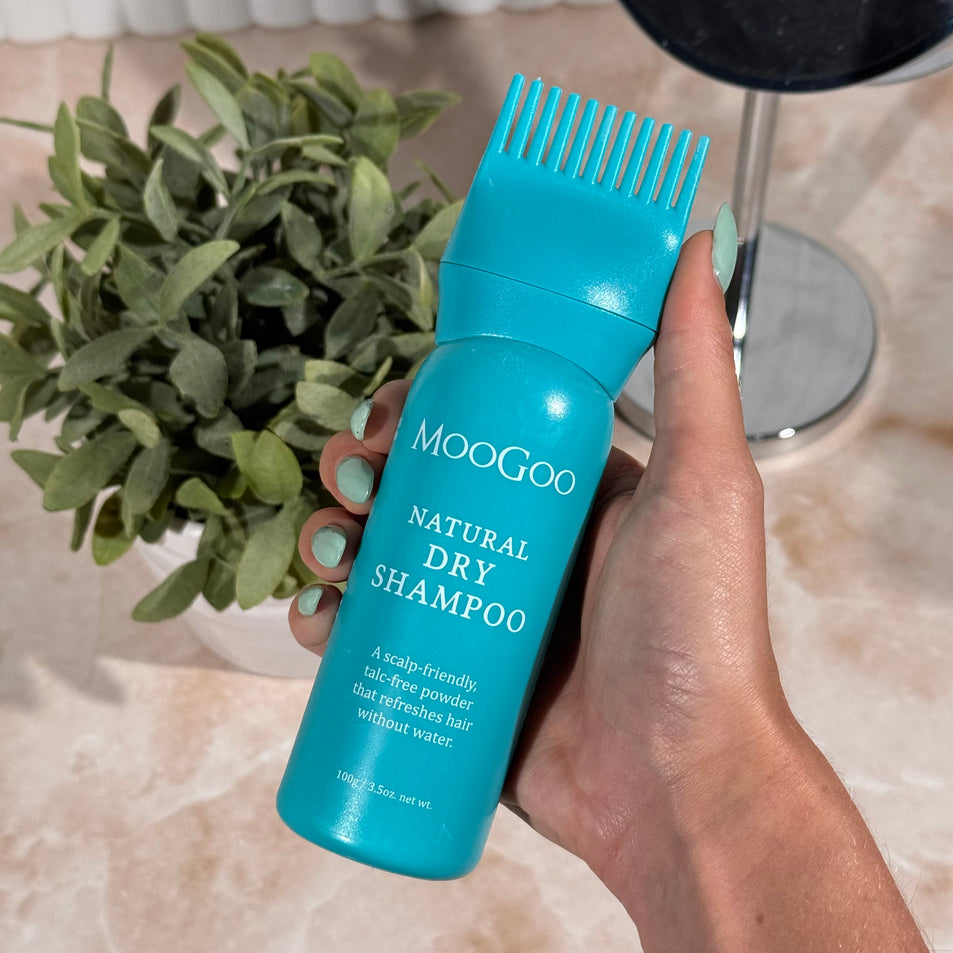 Hand holding a blue bottle of Moogoo Natural Dry Shampoo with a plant in the background. Natural talc free formula. Adds volume and texture without aerosols.